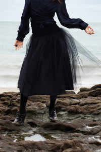 jeune femme brune, vêtue d'un tutu noir lon, et d'un cache coeur foncé à manches longues, les cheveux au vent. Elle est sur un rocher sur la plage. Création Maureen Vinot