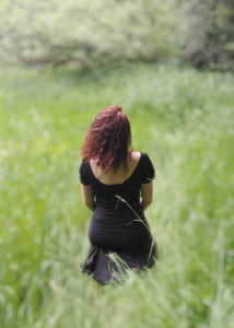femme assise dans une prairie verdoyante, habillée avec un tee shirt dos nu noir et une jupe noire. Création Maureen Vinot