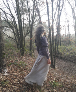femme qui marche dans la forêt et habillée d'une longue jupe de lin fluide. D'un pull beige à manches bouffantes. Ces vêtements sont ceux de la créatrice Maureen. Ils sont fabriqués en Bretagne